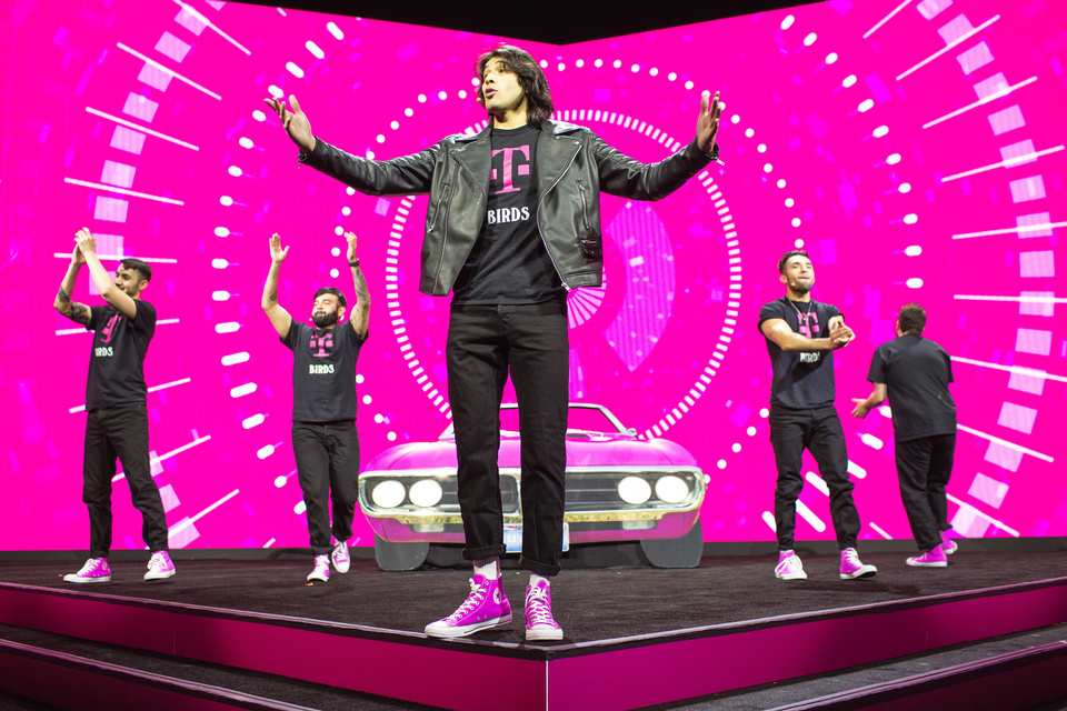 Photo of 5 performers clapping on a stage in front of Magenta set with a magenta car. Performers' shirts read "T-Mobile Birds."
