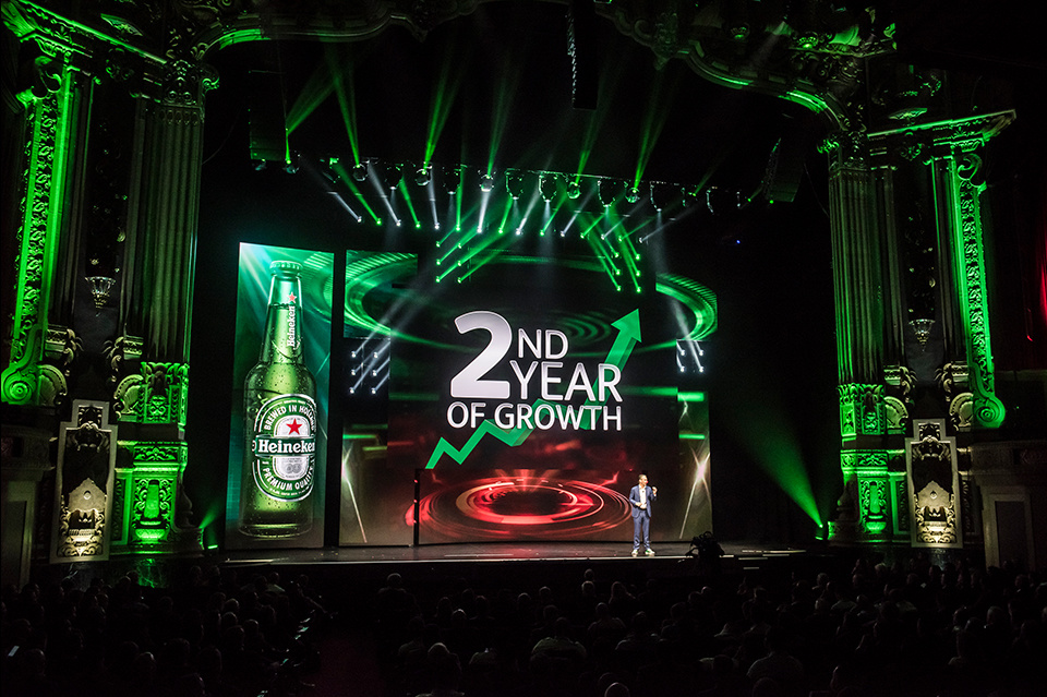 Photo of a presenter on green lit stage addressing a crowd in front of a screen displaying a Heineken bottle and the text "Second year of growth"