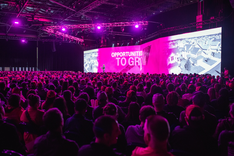 Photo of a large audience watching a presenter on stage in front of a magenta screen that reads "opportunities to grow."