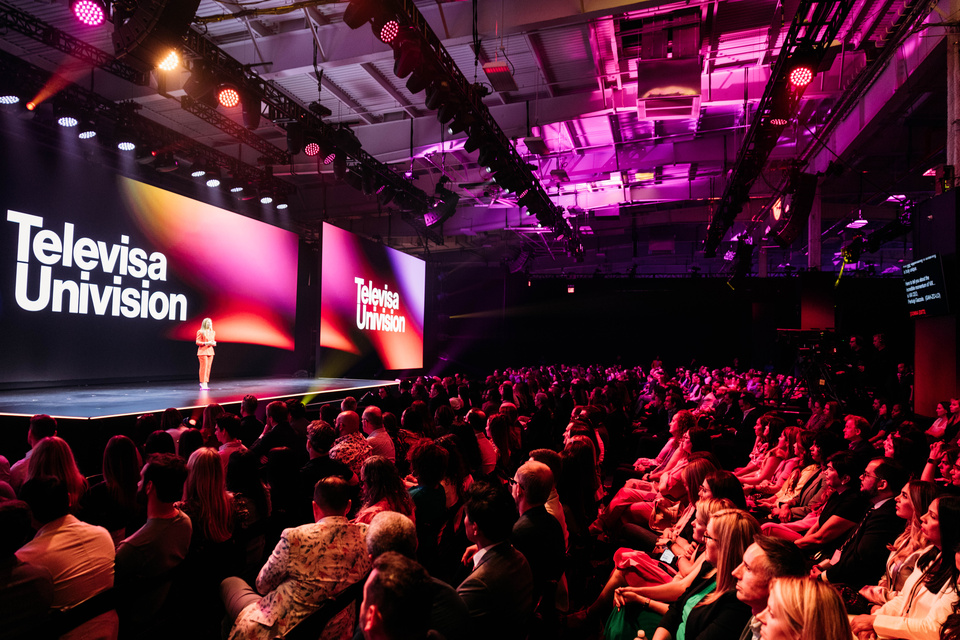 Photo of a Side view of Triple Screen Stage with a Speaker addressing an audience. Televisa Univision branding is on the screens.