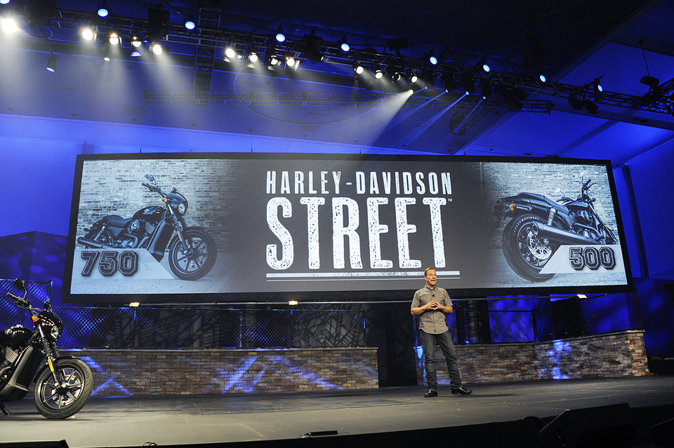 Photo of presenter on stage next to a motorcycle in front of a screen that reads: "Harley-Davidson Street"