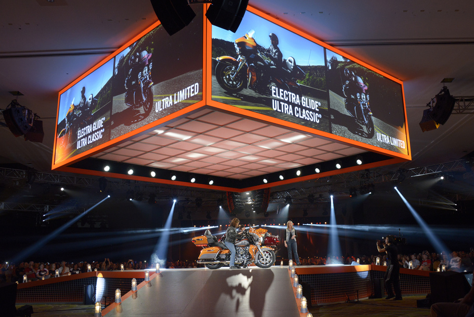 Photo of a person sitting on an orange motorcycle on stage with an orange arena screen showing motorcycle features