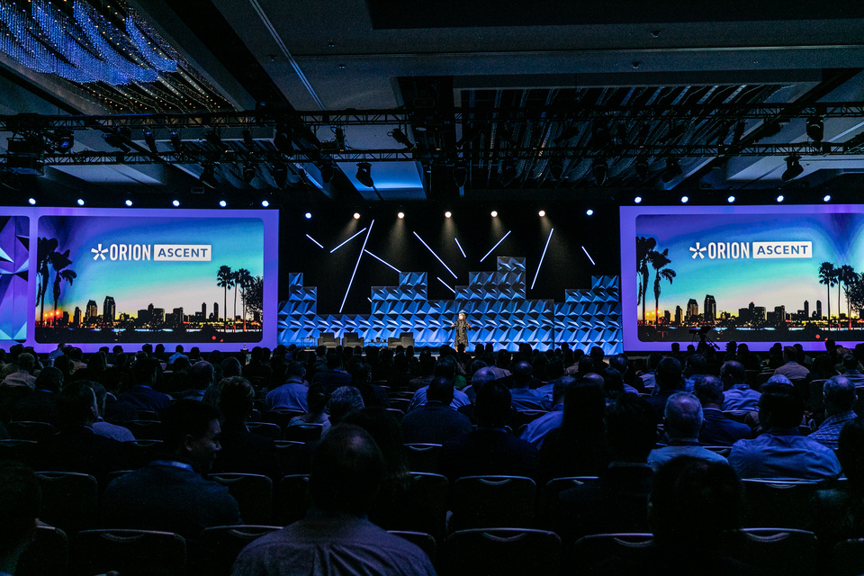 Photo of a host wearing a cape on stage in front of a screen with palm tree graphics