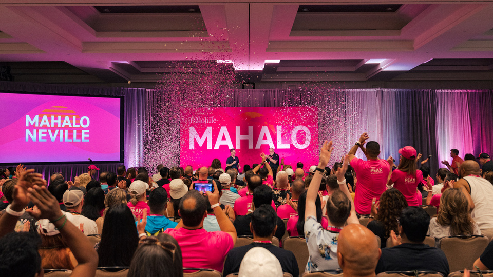 Photo of attendees watching 2 speakers on stage at a town hall with magenta confetti and T-Mobile "Mahalo" signage