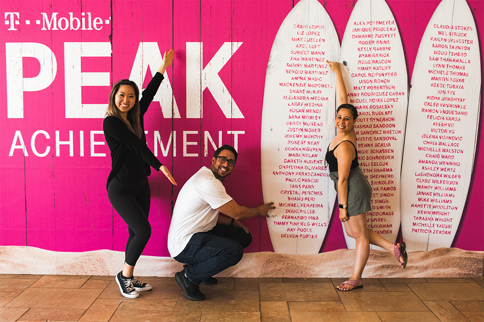 Photo of 3 People pointing at Surf Boards that have a list of Names on them. Magenta branding reads :T-Mobile PEAK Achievement."