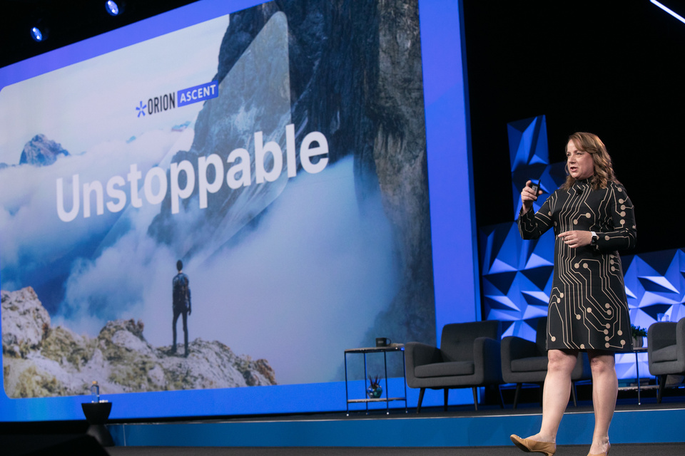 Photo of Orion CEO presenting on a stage in front of a mountain image that reads "Unstoppable" with blue lighting