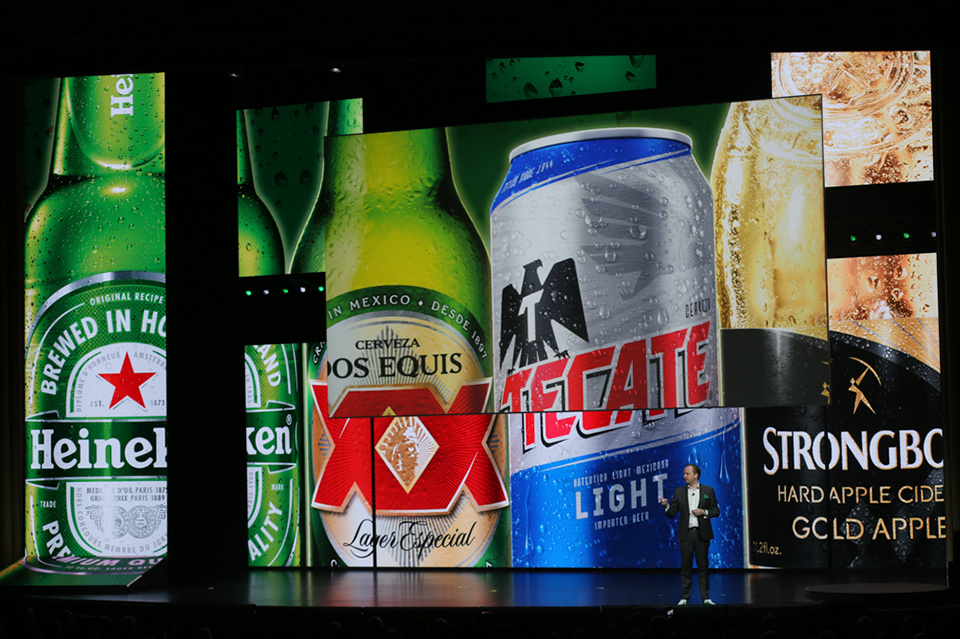 Photo of presenter on stage in front of an LED screen showing Heineken, Dos Equis XX, Tecate and Strongbow cans/bottles