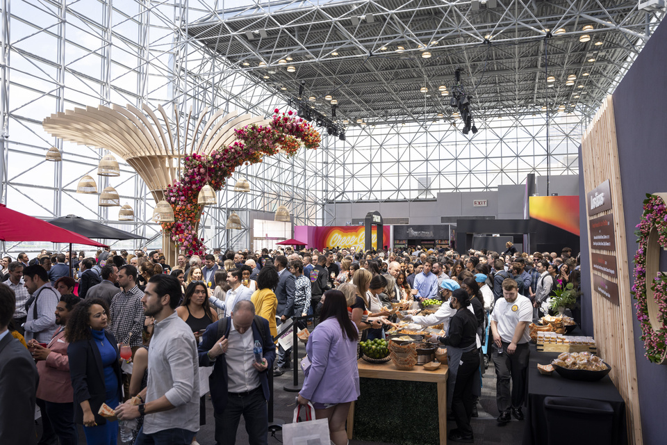 Large crowd at an indoor TelevisaUnivision event with decorative floral displays and food being served, under a glass-paneled ceiling at the Javitz Center in New York.