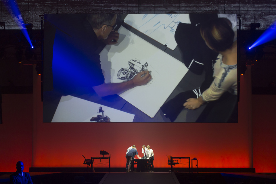 Photo of 4 artists on a stage. 1 artist is sketching a motorcycle.