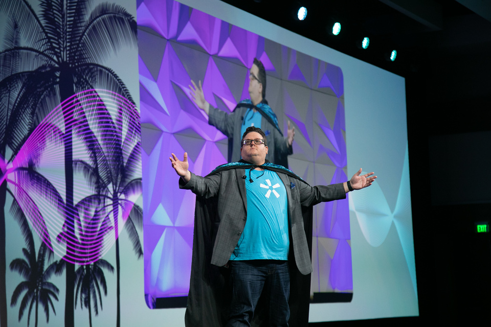 Photo of a host wearing a cape on stage in front of a screen with palm tree graphics
