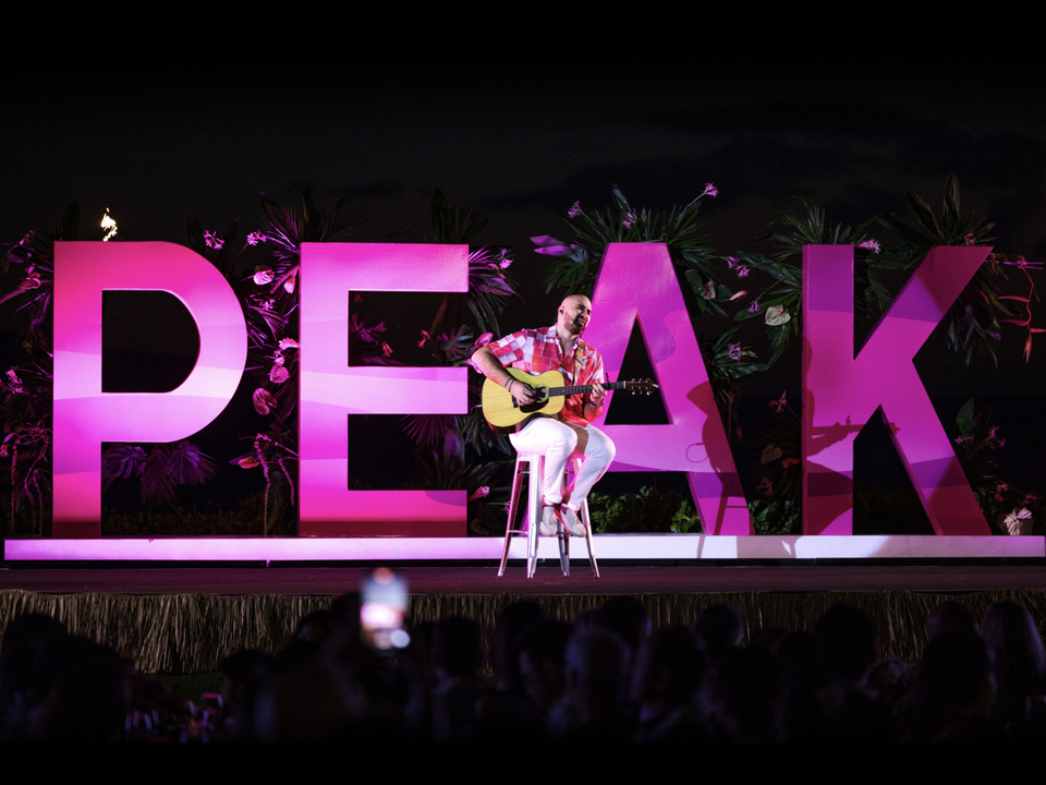 Photo of a musician seated on a stage playing an acoustic guitar in front of large magenta letters that read "PEAK"