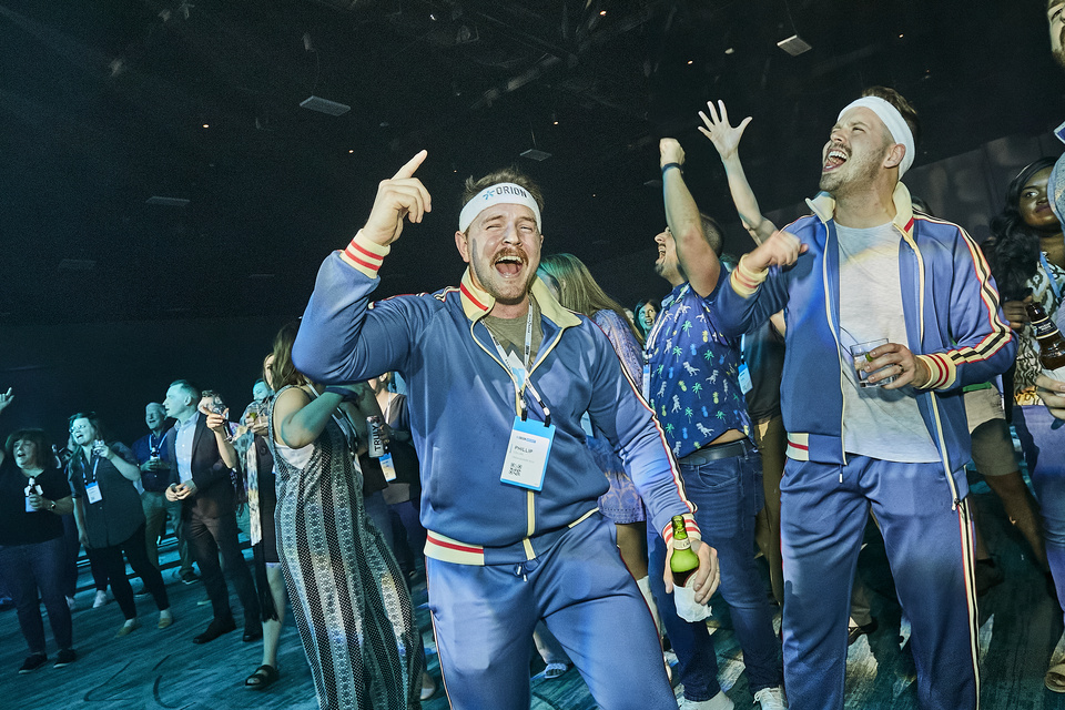 Photo of concert attendees in a crowd, with attendees in the foreground wearing tracksuits with their hands up, singing, and holding drinks