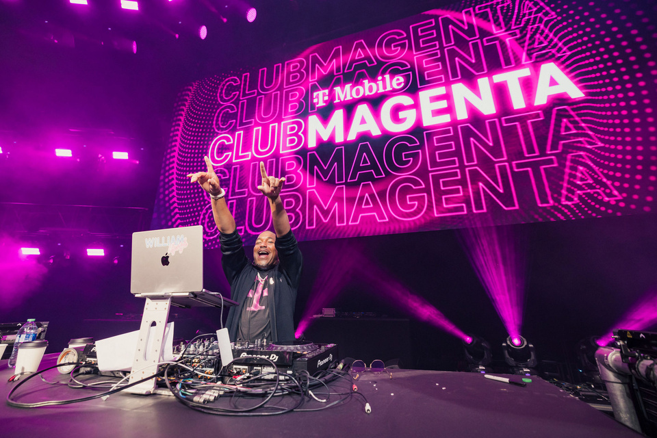 Image of DJ in front of T-Mobile Club Magenta branding. DJ is Smiling and pointing to the audience.