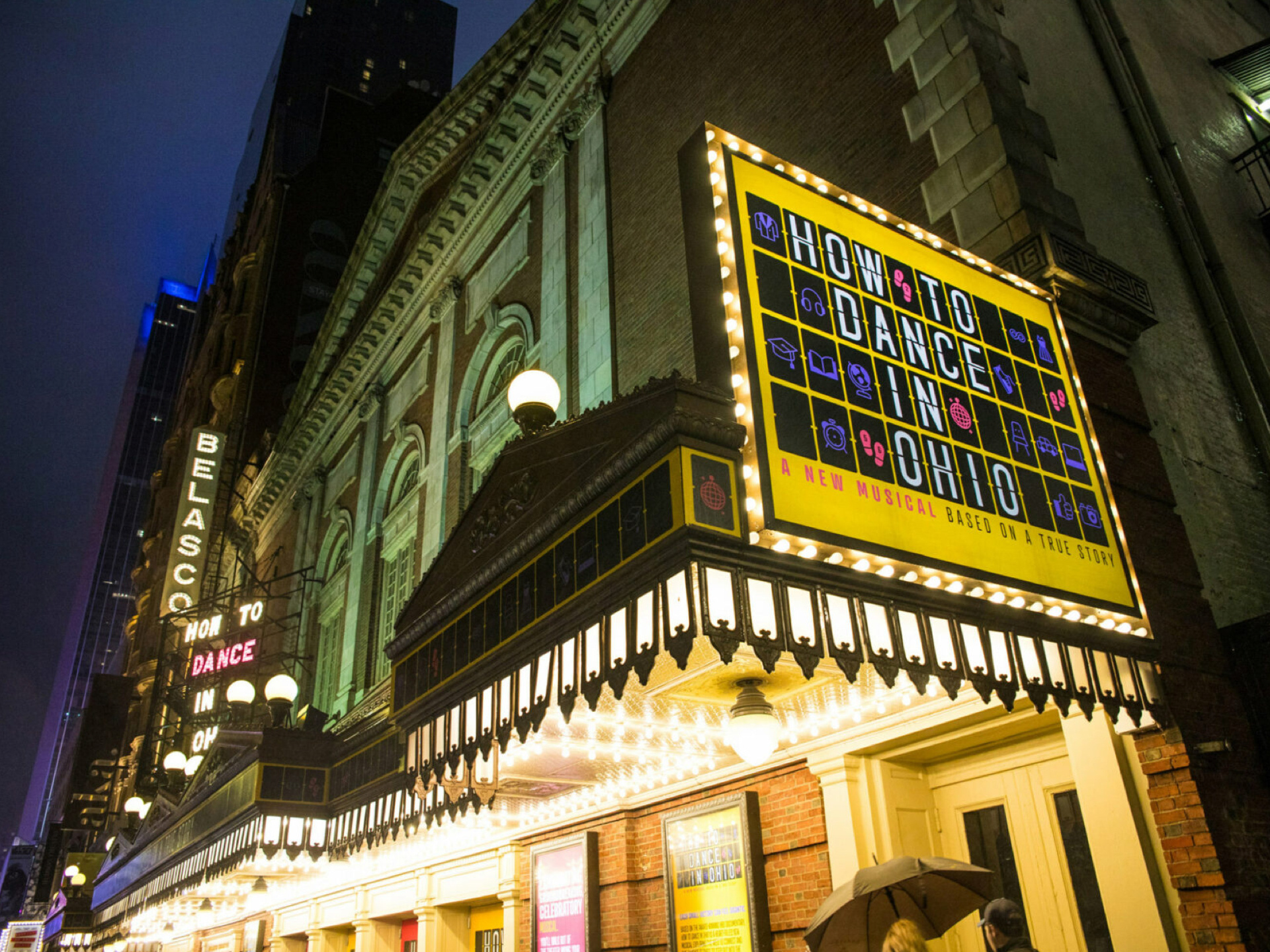 Image of a lit Broadway marquee from the street with a "How to Dance in Ohio: A new musical based on a true story" sign on a yellow backdrop