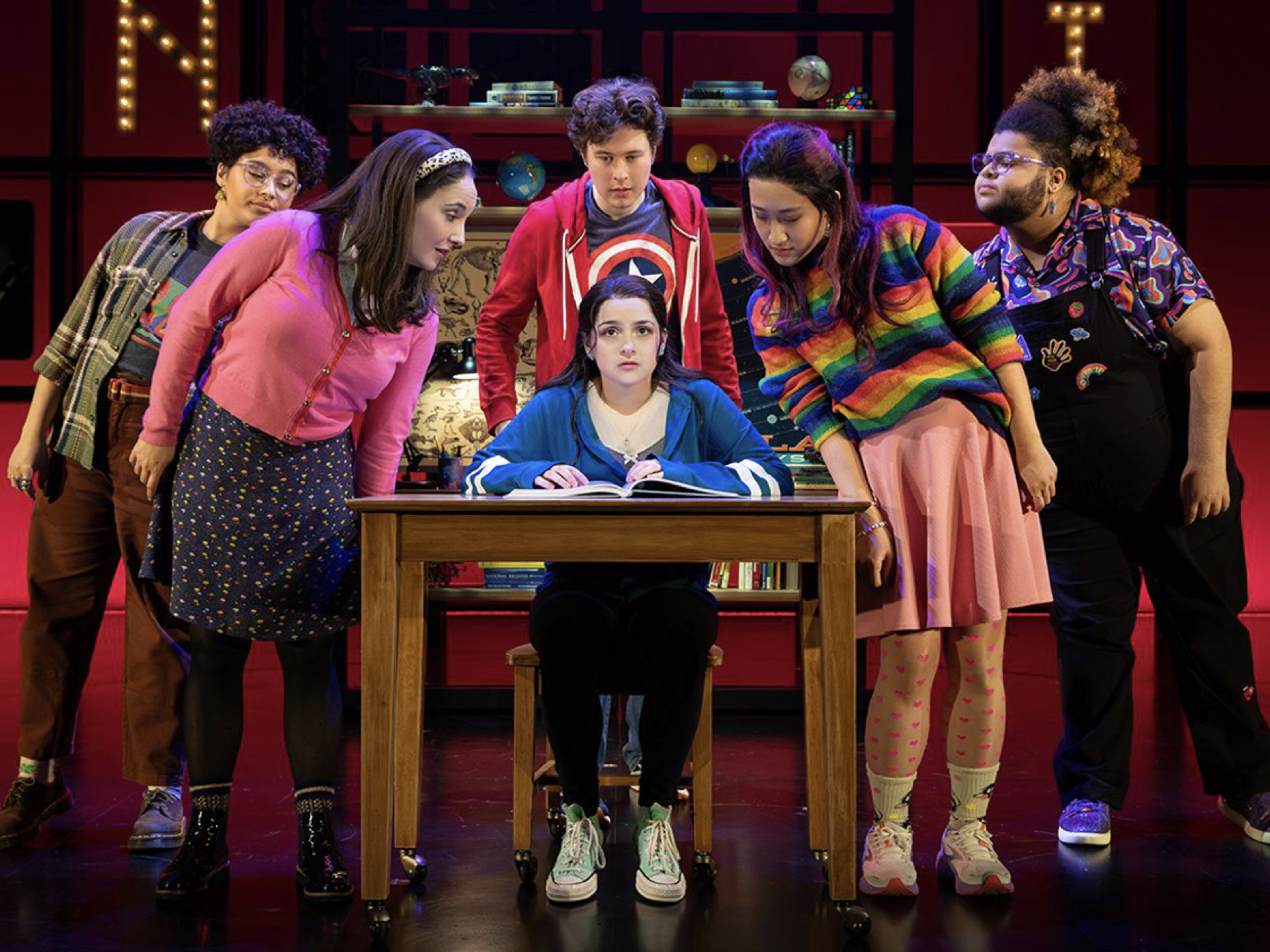 image of actors on a stage with 1 person sitting at a desk with a book and 5 other people surrounding and looking at the book on the desk