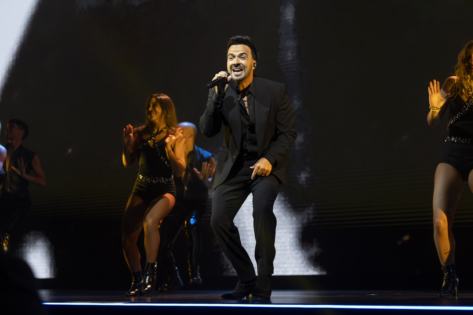 Photo of a male performer Singing into a microphone on a stage in front of multiple Background Dancers