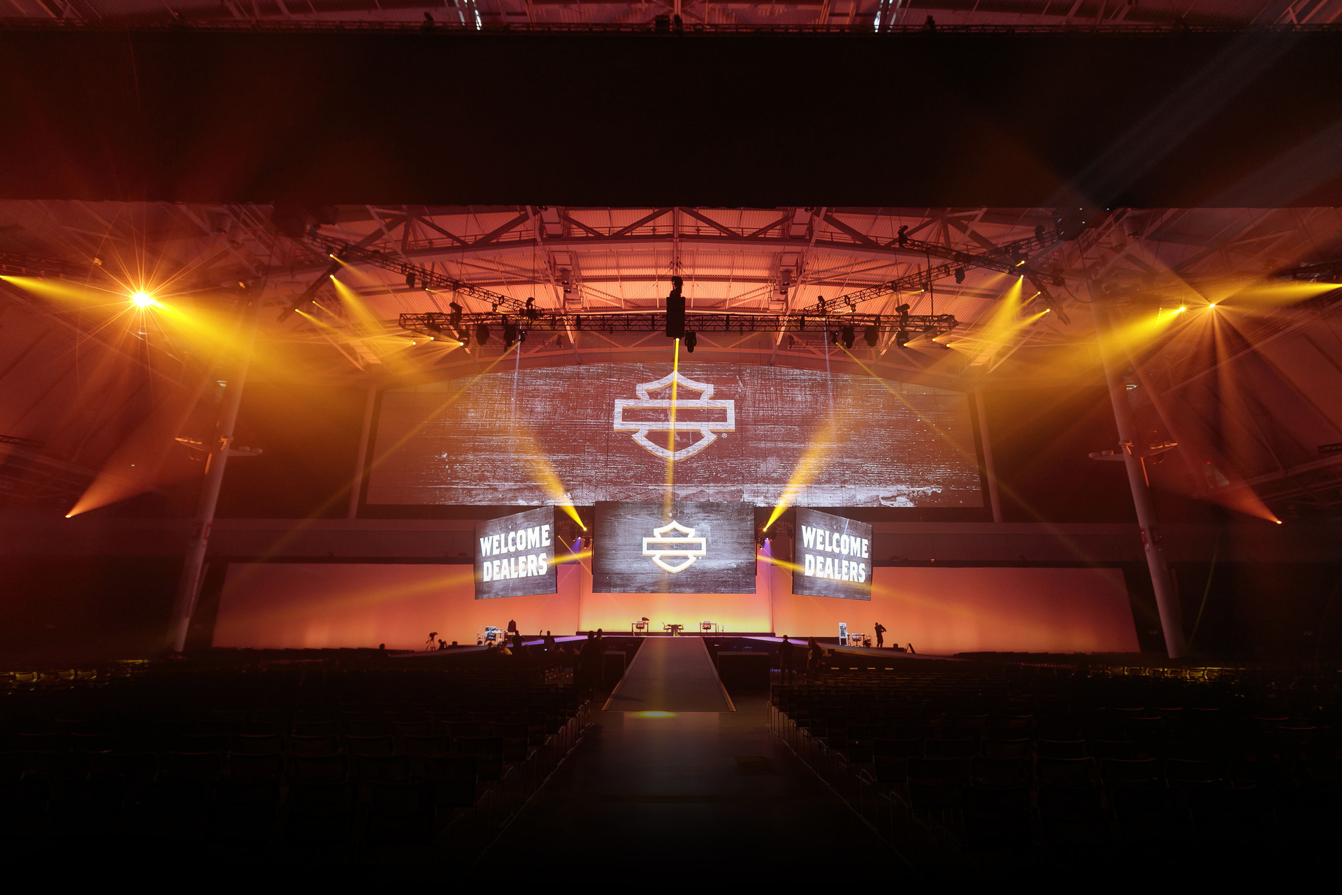 Photo of Harley Davidson event in Boston in orange light. LED screens read Welcome Dealers with Harley Davidson Bar and shield. Room and stage are empty