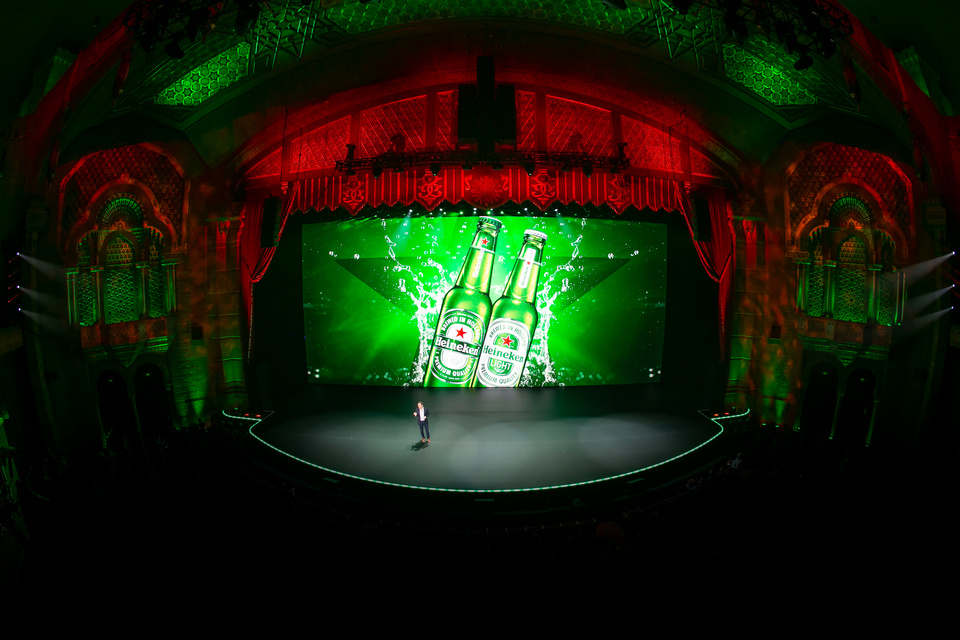 Photo of presenter on stage in a theater in front of an LED screen showing 2 green Heineken bottles.