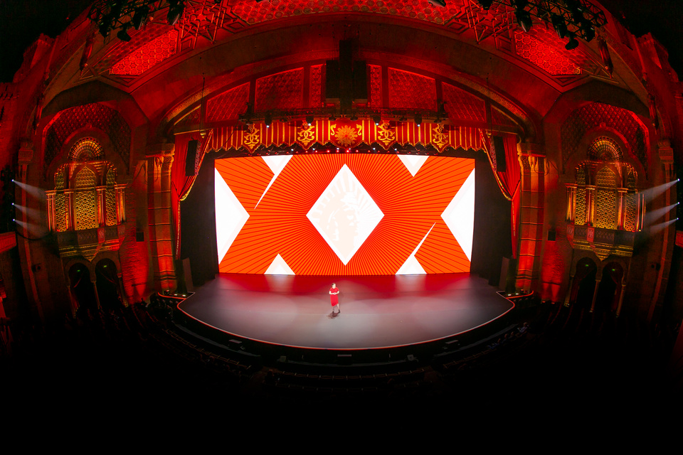 Photo of presenter on stage in a theater in front of an LED screen showing XX logo