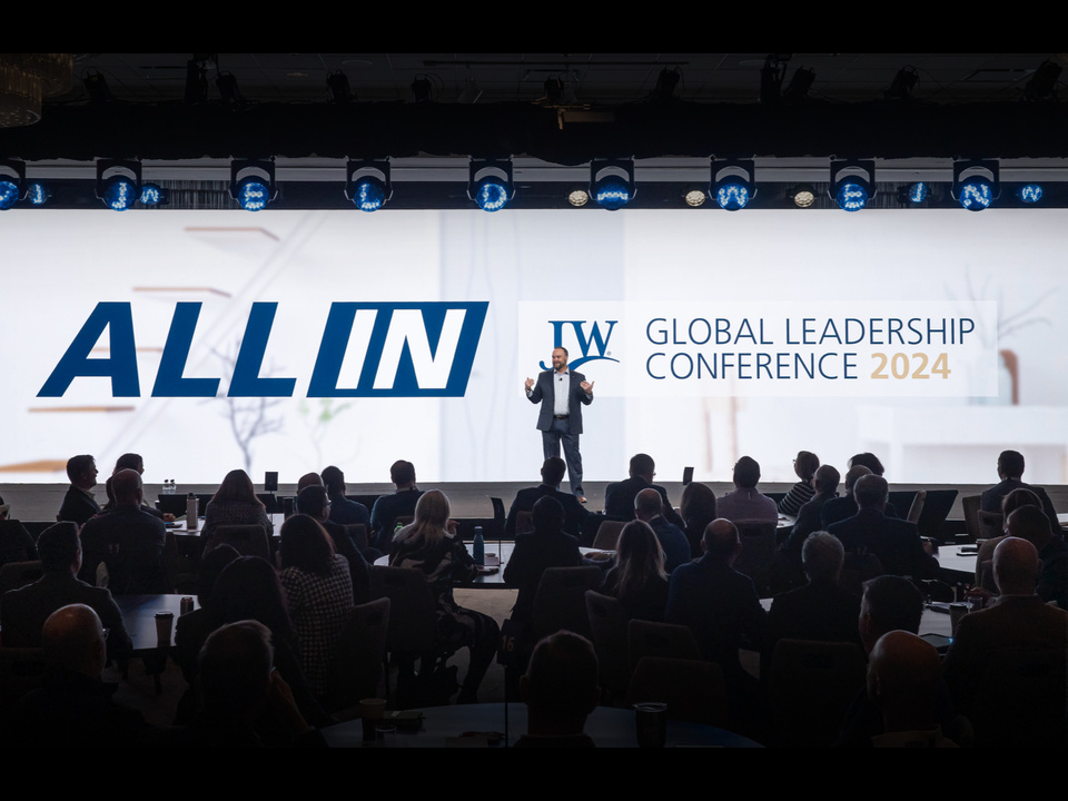 image of a presenter on stage speaking to a group of people sitting at round tables in front of a white background that says "All in" with the JELD-WEN Global Leadership Conference 2024 logo and blue custom JELD-WEN lights