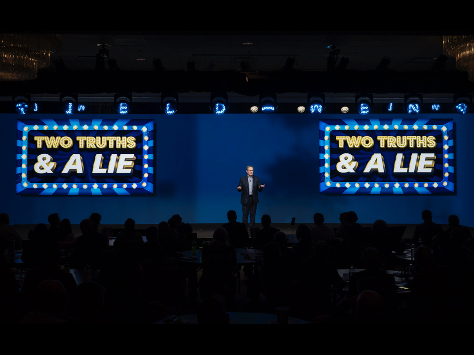 Image of a man in a suit on stage speaking to a crowd in front of a blue background and text on screen that reads "two truths and a lie"