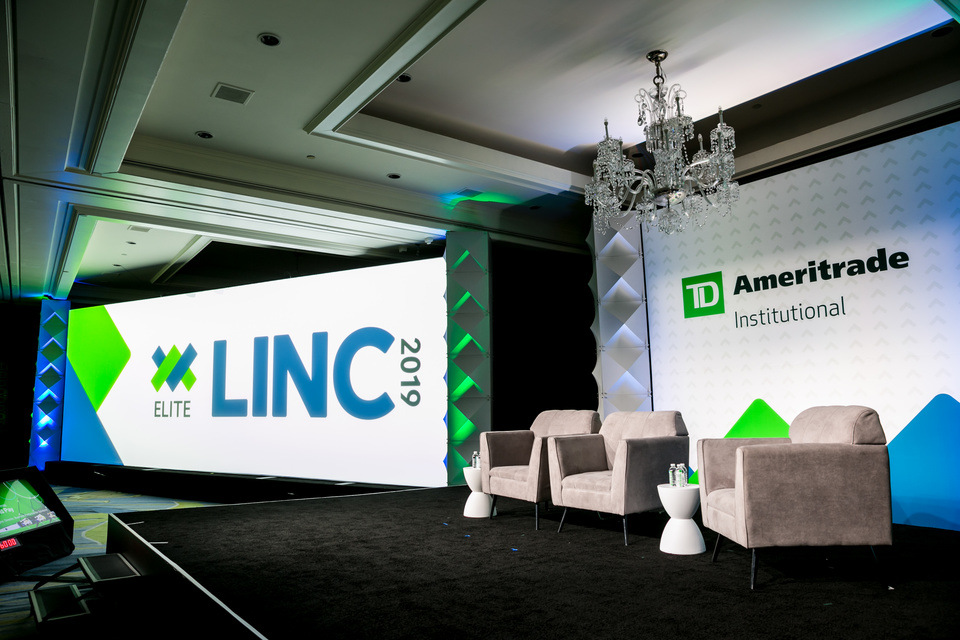 Photo of a small stage with 3 empty tan lounge chairs on the stage. TD Ameritrade Institutional Elite LINC 2019 branding is displayed on 2 screens behind the stage.
