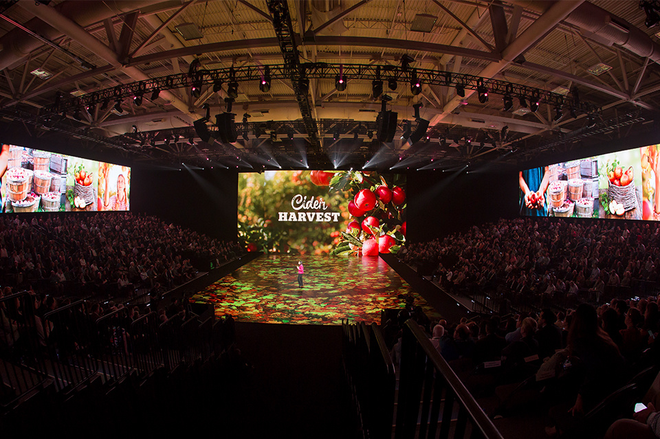 Photo of presenter on apple orchard themed LED stage speaking to an audience. Screen reads "Cider Harvest."