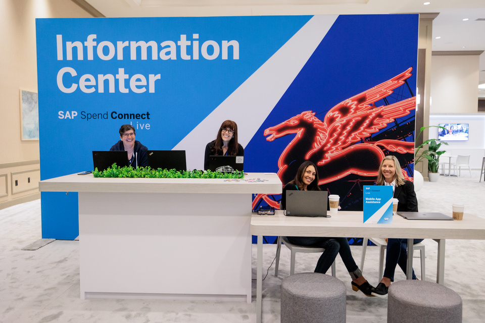 Photo of 4 staff members sitting at desks with laptops in front of an SAP Spend Connect Live Information Center sign