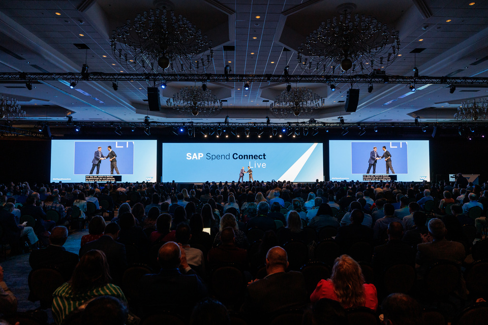 Photo of 100s of audience members watching two people shake hands on stage in front of LED screens that read "SAP Spend Connect Live