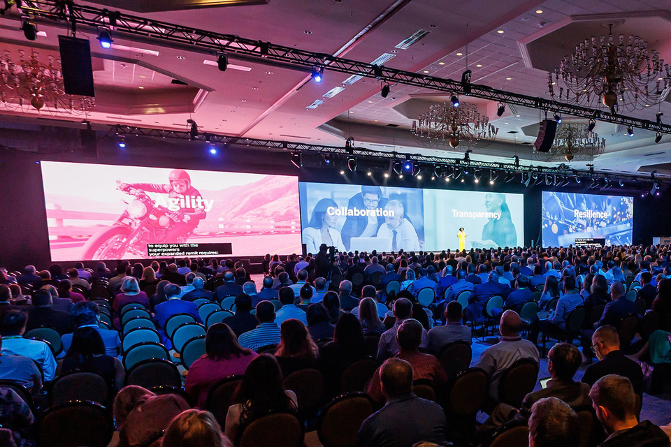 Photo of 1 person delivering a presentation to a crowd of people in front of 4 screens that read "agility, collaboration, transparency, and resilience"