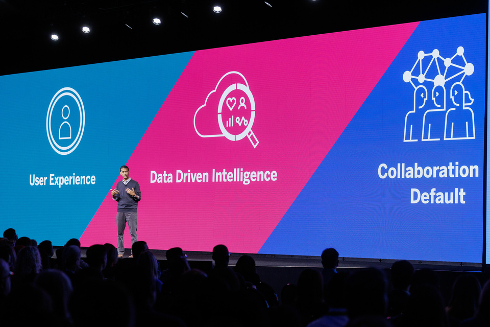 Photo of a presenter on stage speaking to a large audience. An LED screen behind the speaker reads "User experience, data driven intelligence, collaboration default."