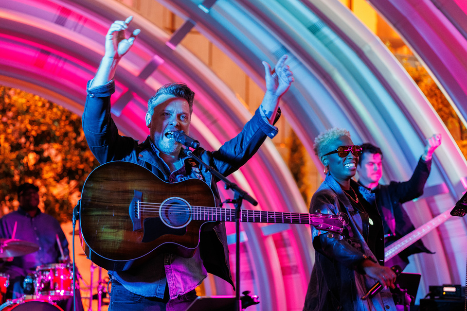 Photo of a performer on stage with an acoustic guitar singing with his hands in the air. 3 band members are shown out of focus behind the guitarist against a colorful background