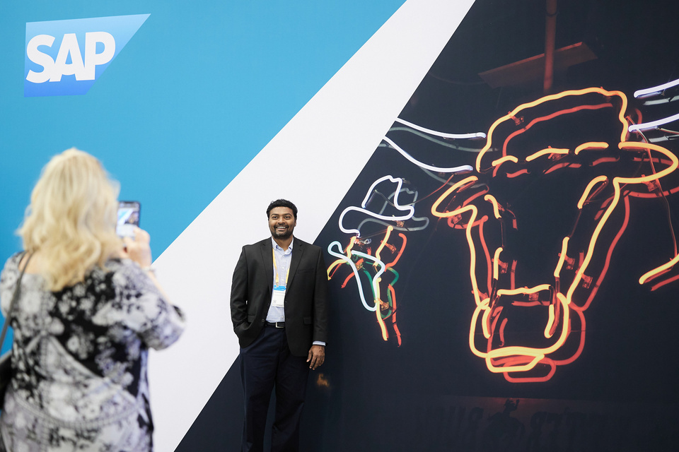 Photo of a woman taking a photo with her phone of a man standing in front of an SAP photo-op that displays a neon bull and cowboy graphic.