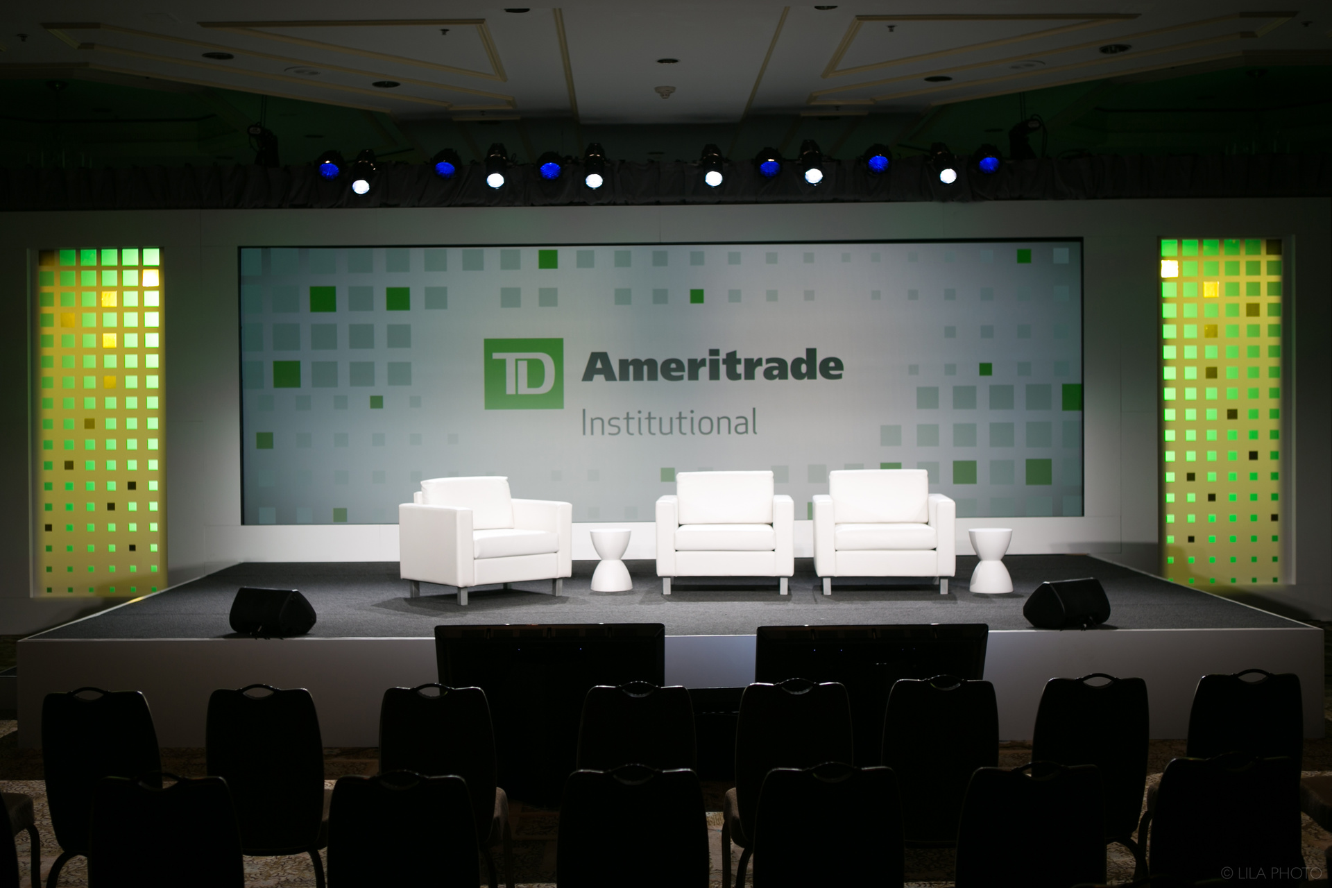 Photo of an empty TD Ameritrade Institutional stage with green lighting. 3 white chairs are set on stage with 2 rows of empty audience chairs
