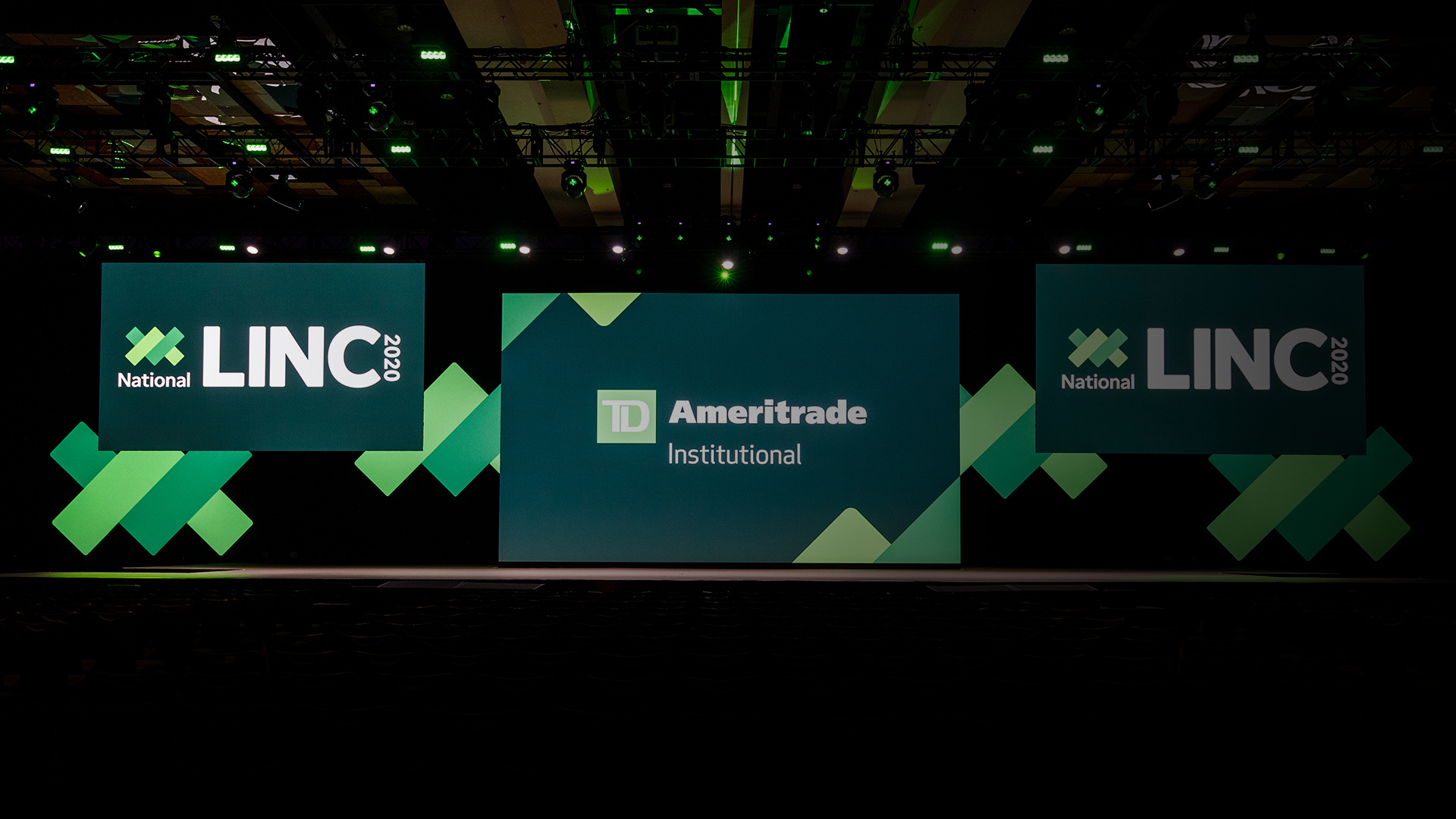Photo of an empty stage with 3 screens showing National LINC 2020: TD Ameritrade Institutional branding and green lighting
