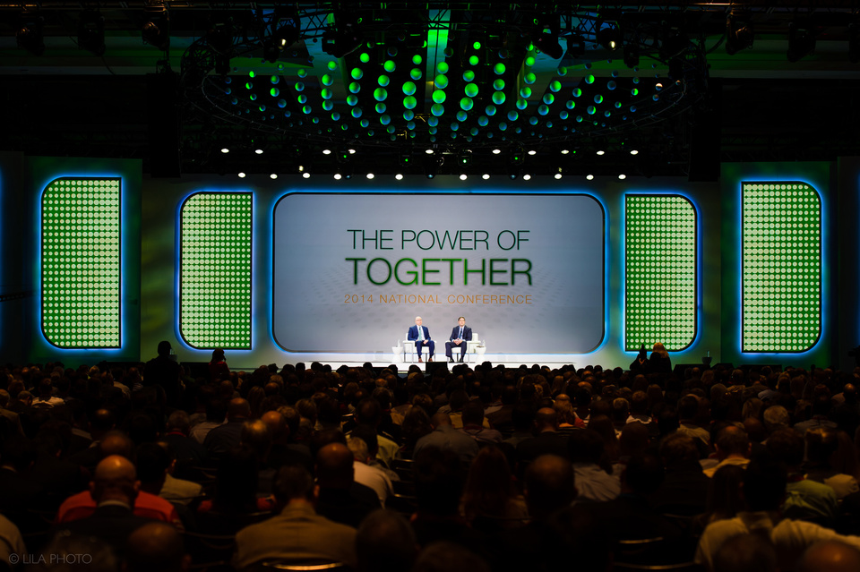 Photo of 2 men seated in white chairs on a stage in front of a large audience. Stage is lit green with a screen that reads "The Power of Together: 2014 National Conference"