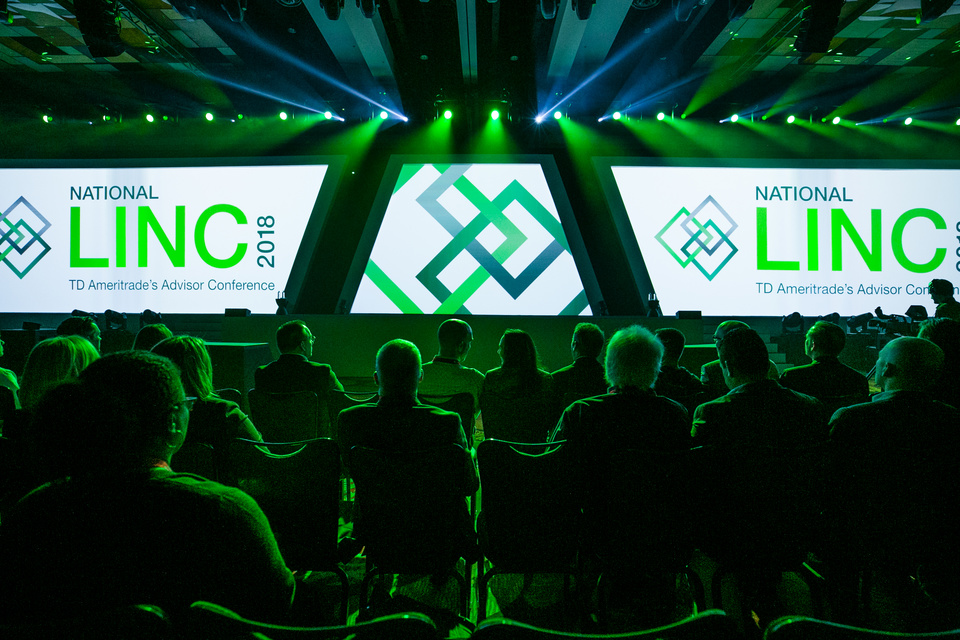Photo of audience members looking onto an empty stage with TD Ameritrade's Advisor Conference National LINC 2018 displayed on Three Screens with green lighting.
