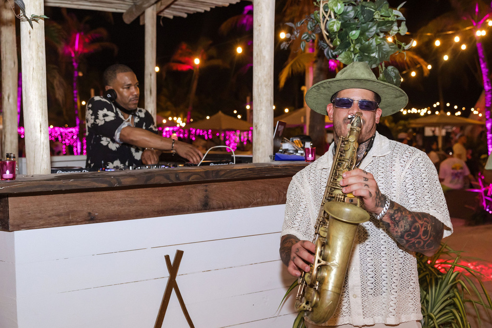 Photo of a saxophonist playing at an outdoor beach venue. DJ is in the background spinning in a white DJ booth