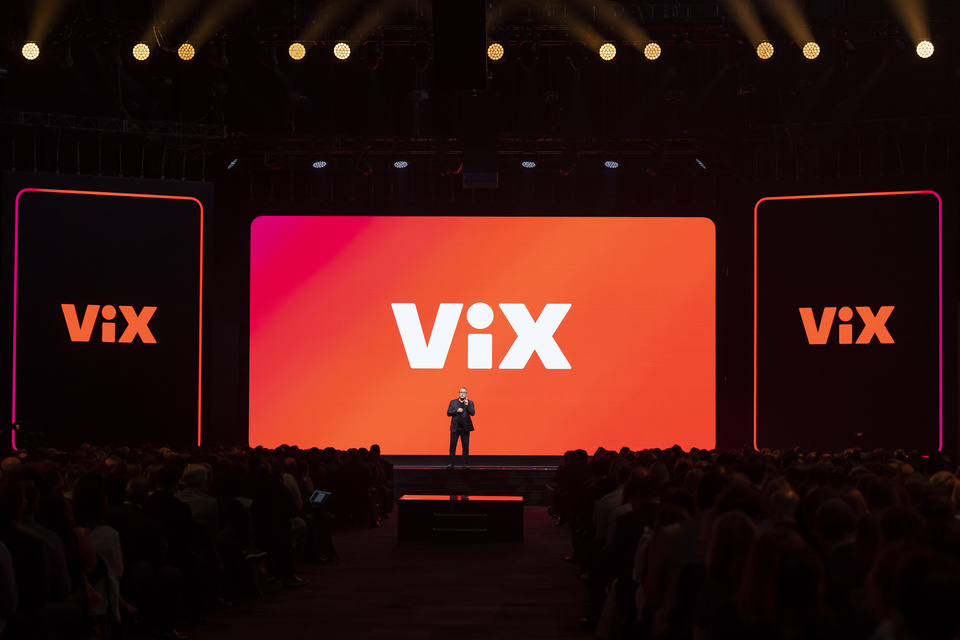 Photo of a presenter on stage in front of an orange screen that reads "Vix"