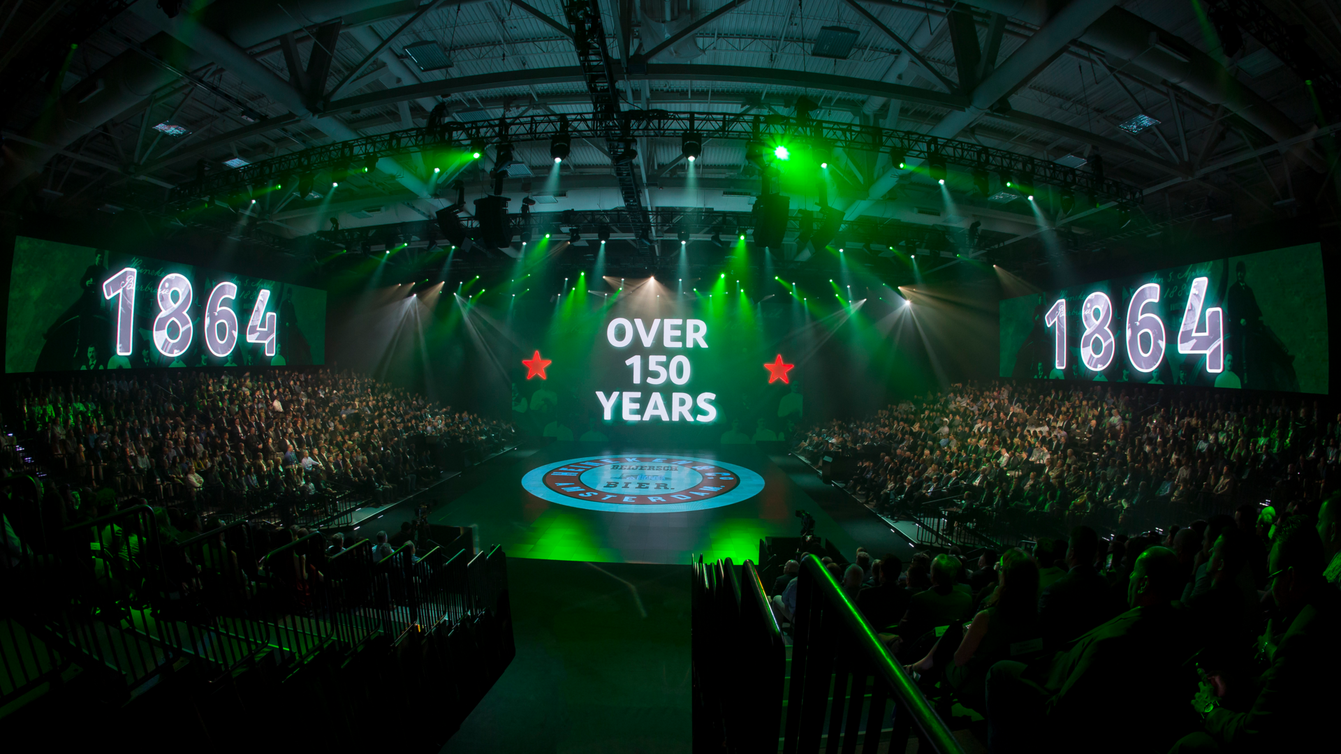 Photo of Heineken stage with guests in the audience on all sides of the stage. LED screens read "Over 150 Years, 1864" in green lighting