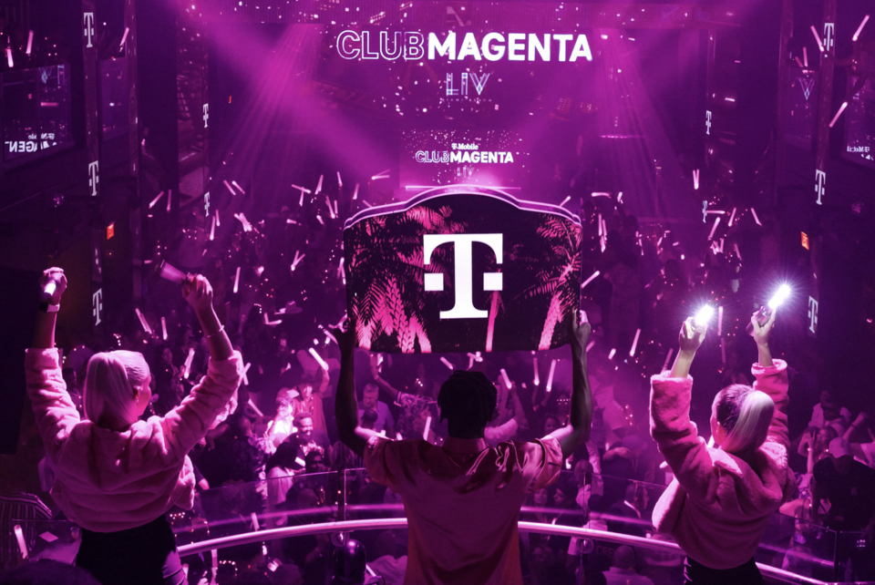 Photo of bottle service staff holding a T-Mobile sign in a crowded magenta lit club with T-Mobile Club Magenta on a screen in the background