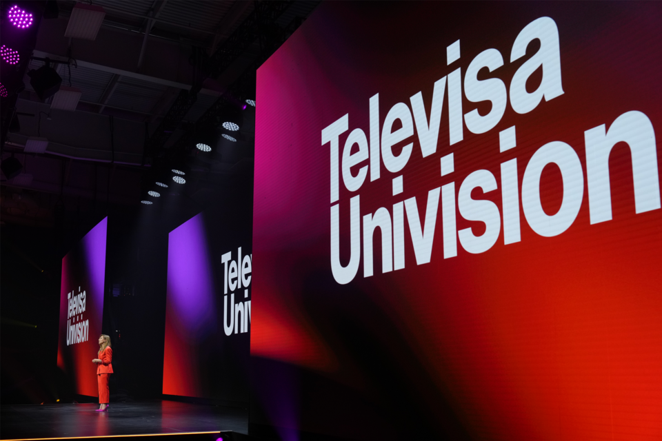 Photo of a Side view of Triple Screen Stage with a Speaker addressing an audience. Televisa Univision branding is on the screens.