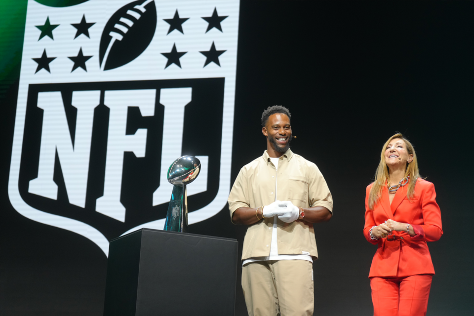 Photo of 2 people smiling on stage next to football trophy with NFL branding on the screen behind them.