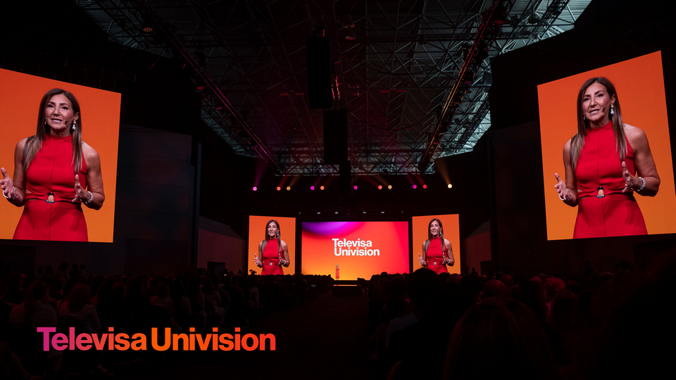 Photo of a Speaker on stage at a TelevisaUnivision Upfront event, with large screens displaying her image and the event logo. Speaker is addressing the audience.