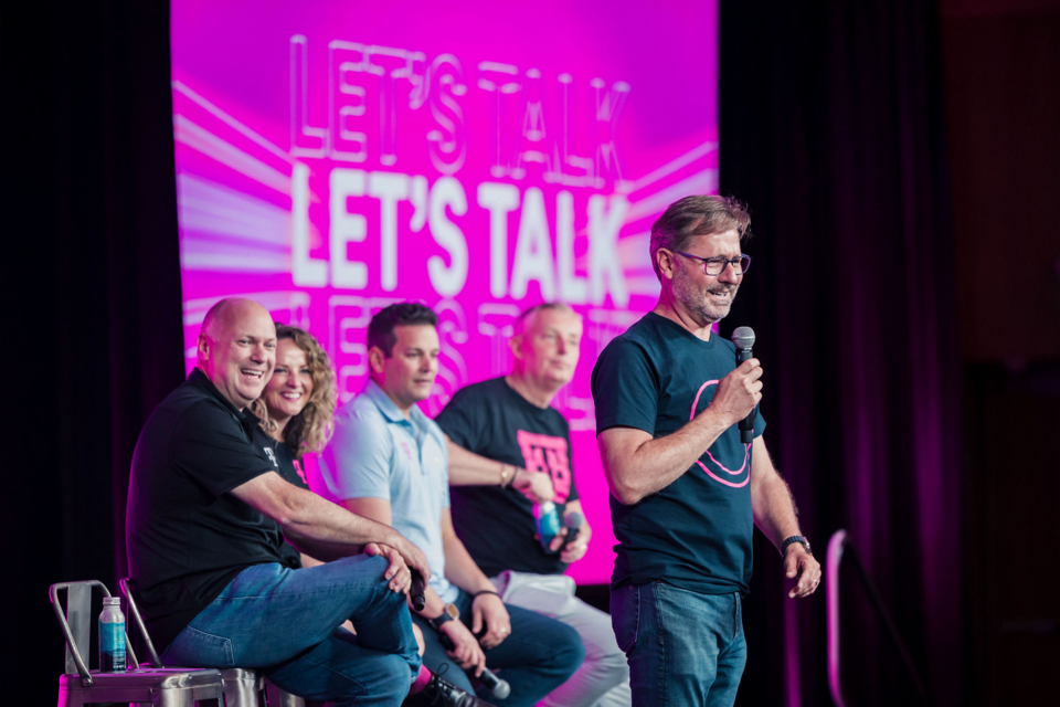 Photo of T-Mobile CEO speaking on stage with 4 other panelists seated and smiling. Signage reads: "Let's Talk"